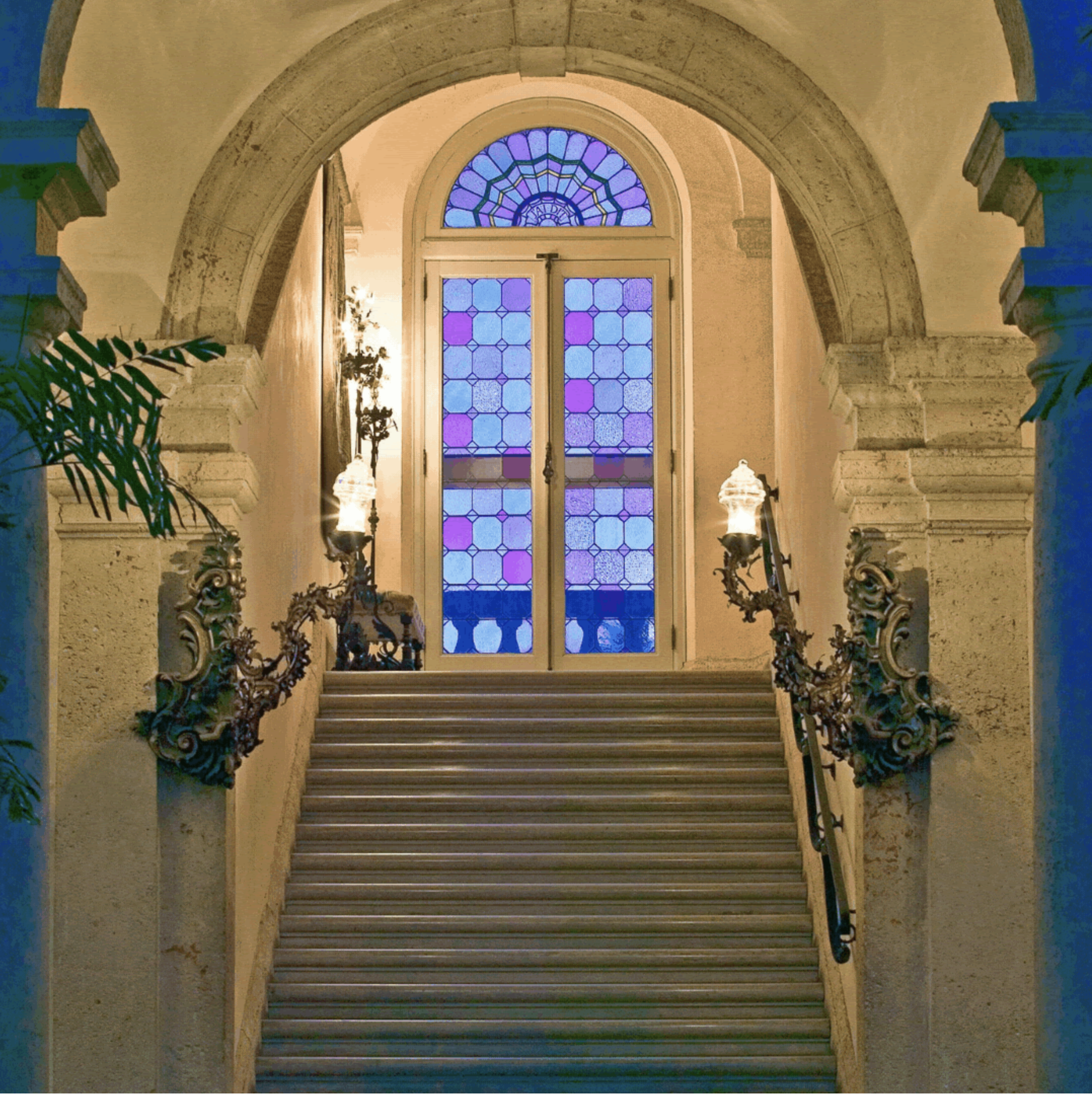 A limestone stairway ascends to a landing with a multi-colored stained glass window, ornate lanterns frame either side